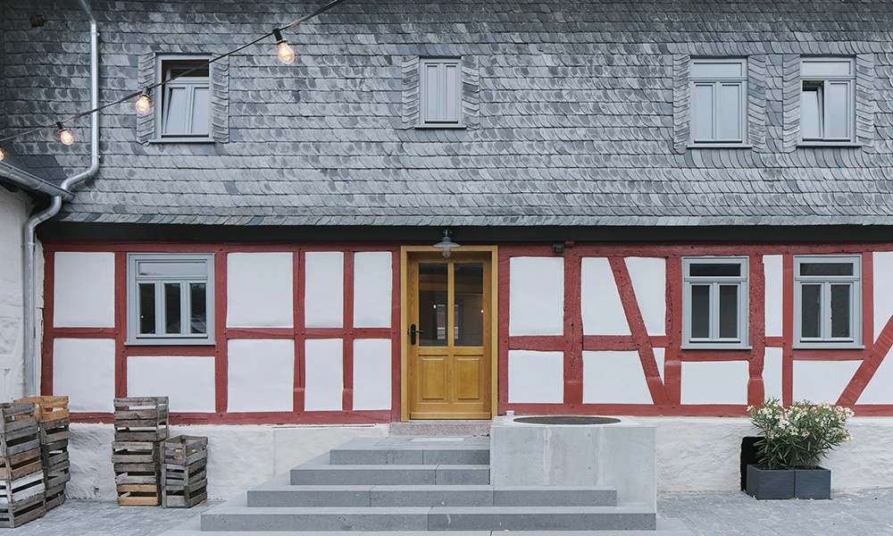 Das Bild zeigt die Fassade eines traditionellen Fachwerkhauses mit roten Balken, weißen Wänden, einem Schieferdach und einem schlichten Eingang mit Holztür und Treppenstufen.