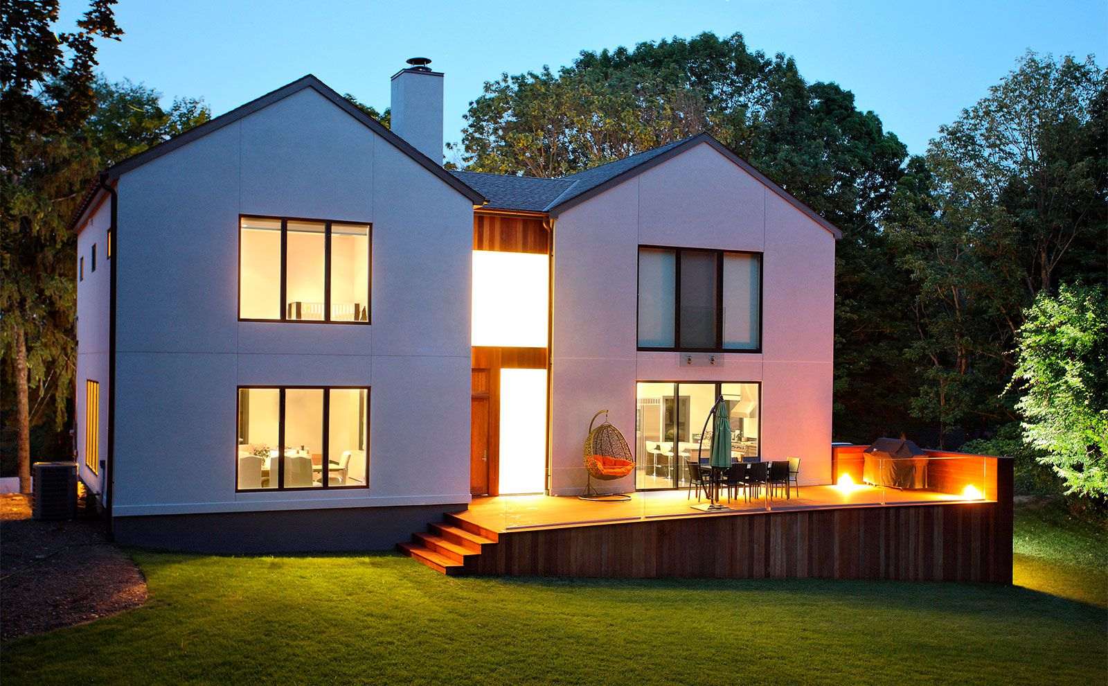 An image of a modern house at dusk, with interior lights on and exterior deck lighting.