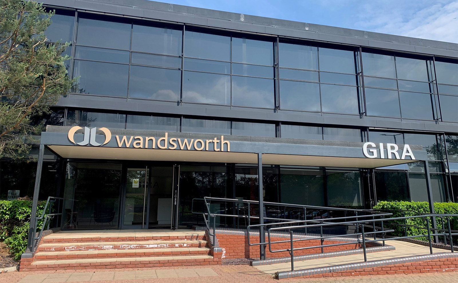 Exterior view of the Gira UK and Wandsworth building, featuring a modern glass facade and a covered entrance with steps and accessibility ramps.