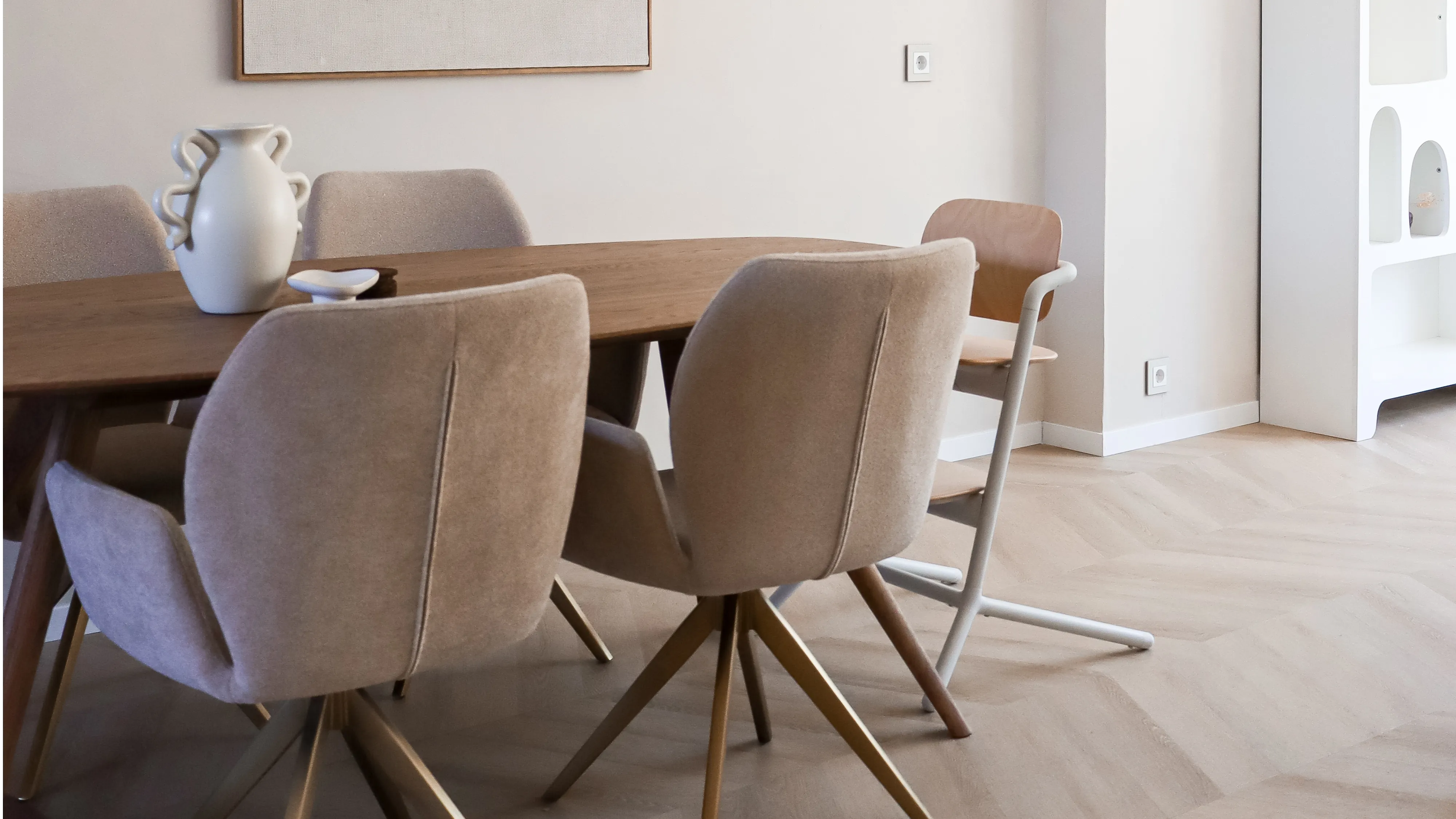 A minimalist dining room with a wooden table, beige chairs, and wall-mounted shelves, featuring subtle wall sockets and switches.