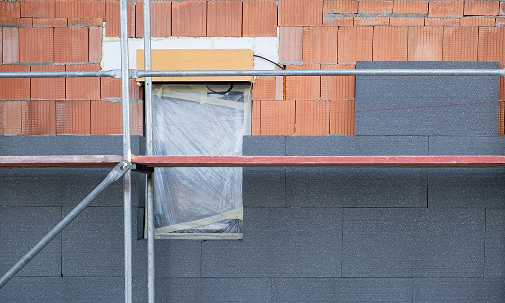 Eine Fassade in der Sanierung: Sichtbare Ziegelsteine werden mit einer neuen Dämmung aus grauen Platten versehen. Ein Gerüst ist für die Arbeiten aufgebaut, während ein Fenster provisorisch mit Folie abgedeckt ist.