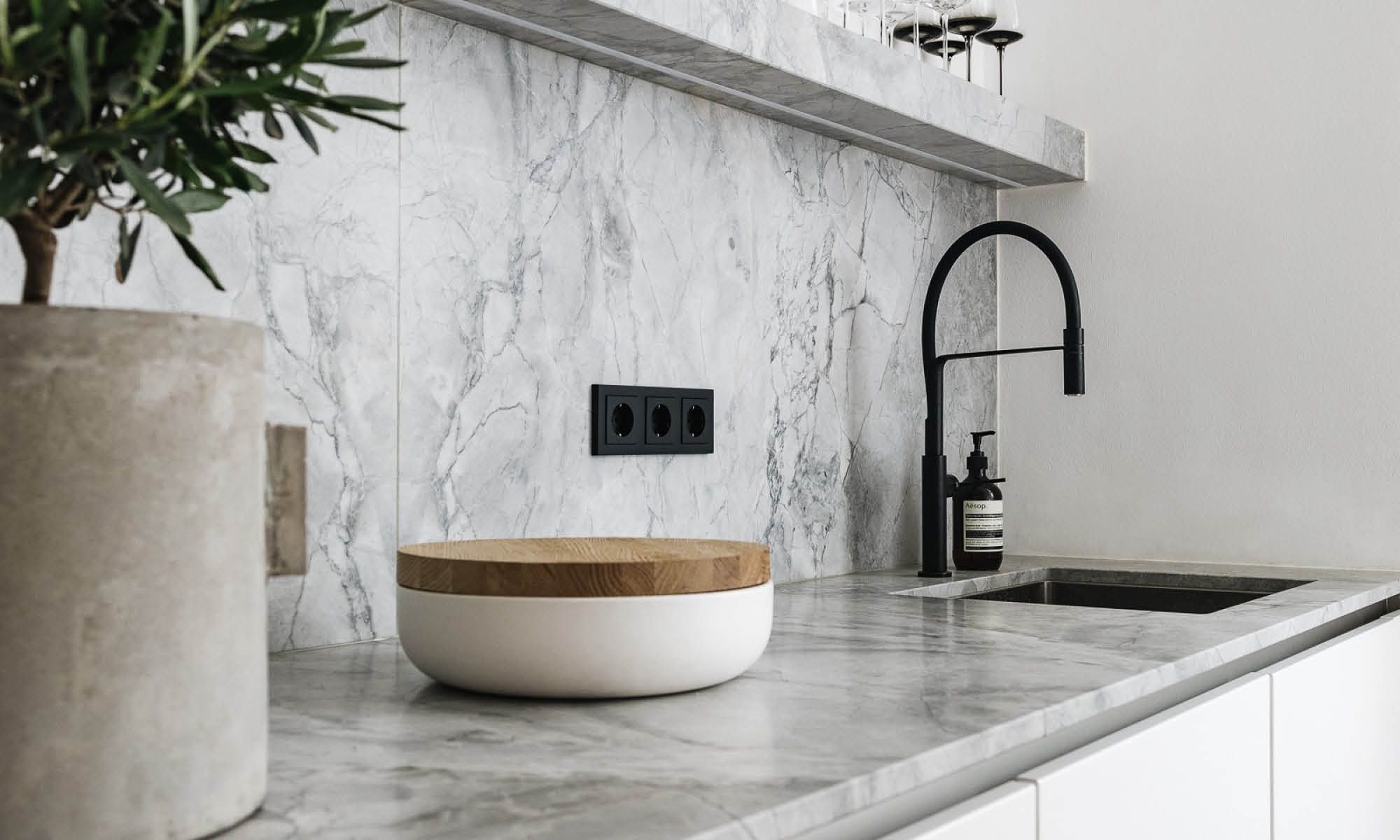 Modern kitchen with a marble countertop and backsplash featuring a black faucet, a white bowl with a wooden lid, and a potted plant.