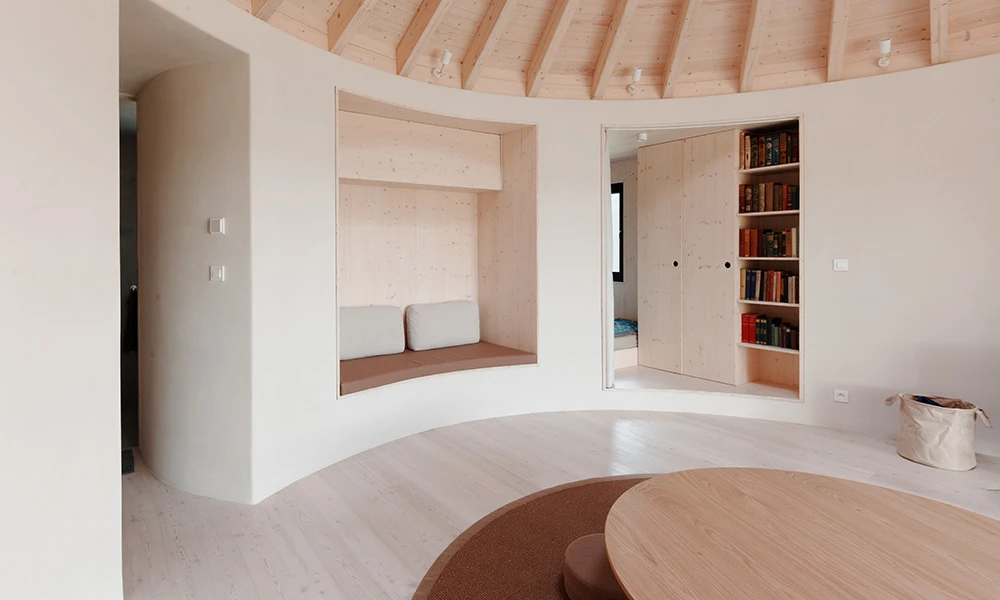 Interior of a modern yurt house featuring a cosy built-in seating nook with cushions, a circular wooden table, and a built-in bookshelf.