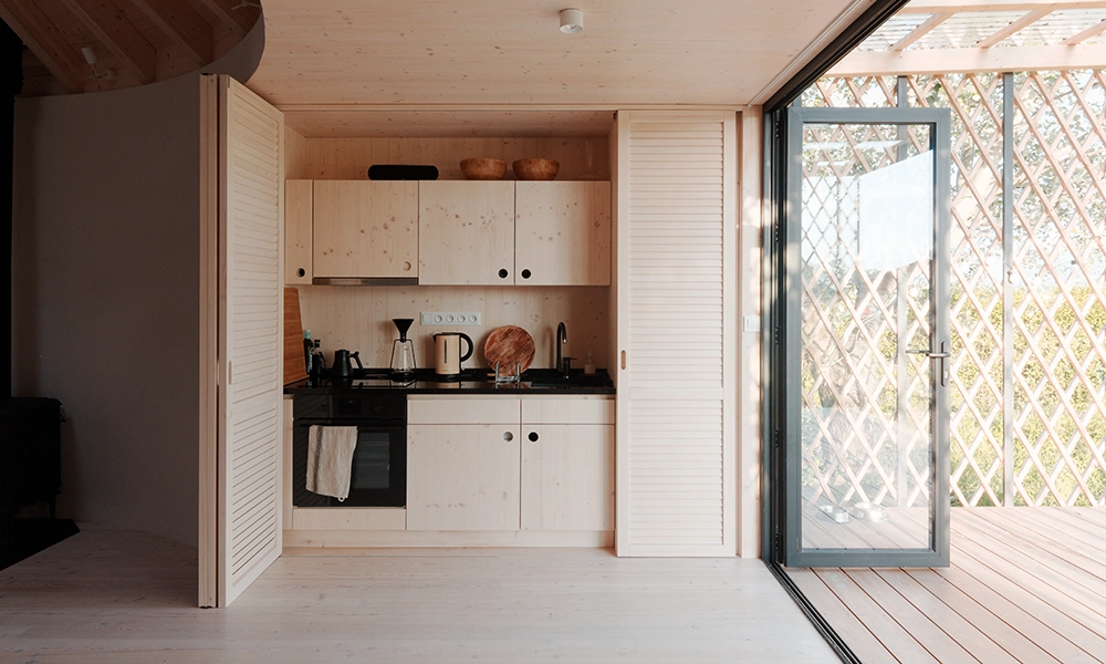 Compact kitchen area in a modern yurt with light wooden cabinets, black countertops, and a sliding glass door leading to an outdoor deck.