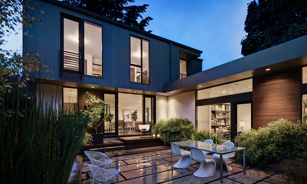 Modern house exterior at dusk with lights on and patio furniture.