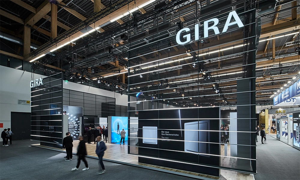 A modernly designed exhibition stand with the "GIRA" logo is visible, surrounded by people passing by.