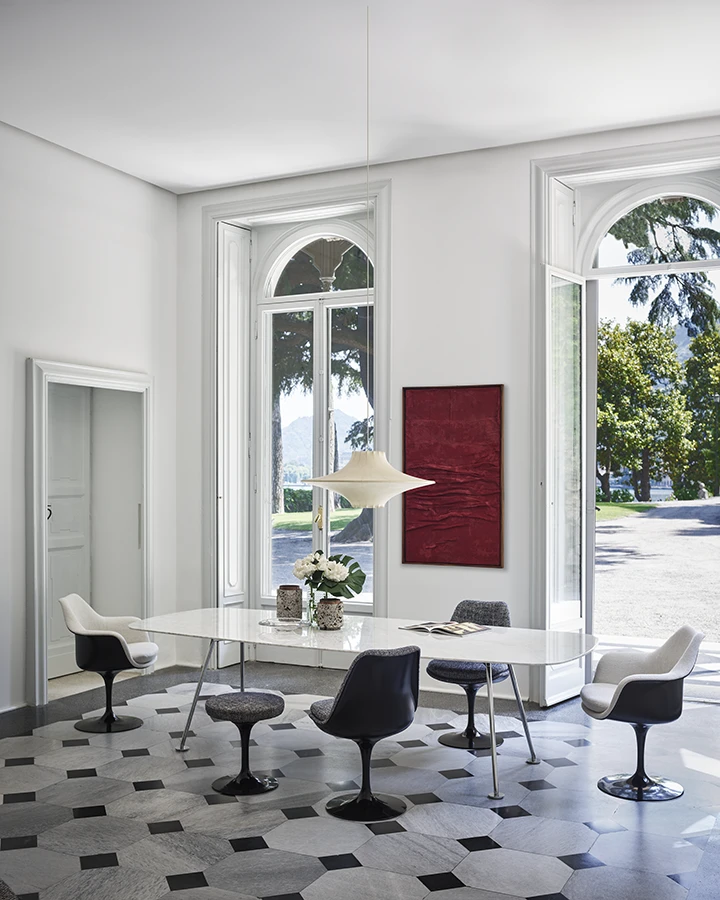 Ein elegantes Wohnzimmer mit modernen Designerstühlen und einem großen, weißen Tisch wird von natürlichem Licht durch bodentiefe Fenster durchflutet, während draußen eine ruhige Parklandschaft zu sehen ist.