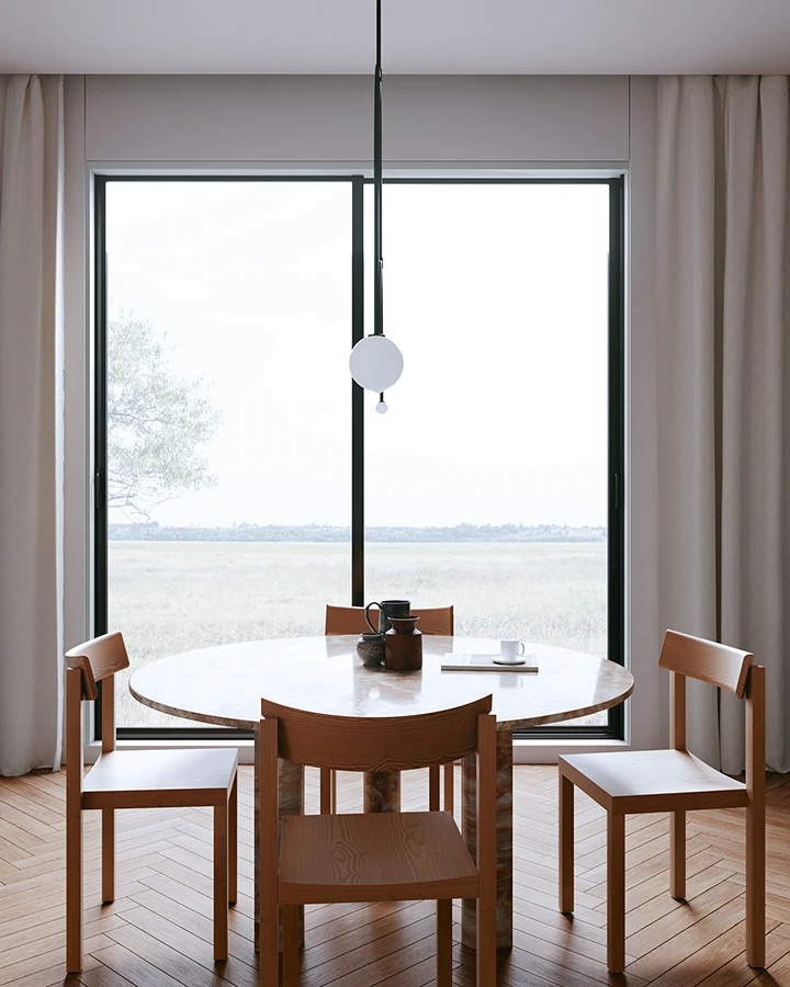 Ein schlichter Essbereich im Mid Century Interior Stil mit einem runden Marmortisch und vier Holzstühlen, der vor einem großen Fenster mit Blick auf eine weite Landschaft steht. 