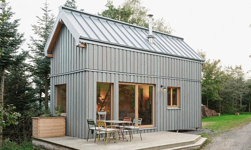 Das kleine, Organic Tiny House mit grauer Holzfassade befindet sich in einer natürlichen, bewaldeten Umgebung.