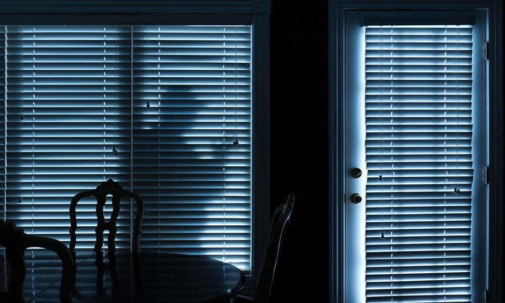 Ein dunkles Zimmer mit einem Esstisch und Stühlen, durch dessen Jalousien man eine schemenhafte Gestalt im Licht draußen erkennt.