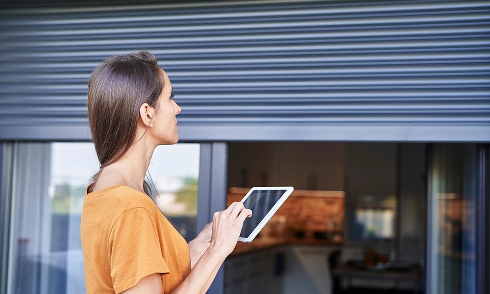 Das Bild zeigt eine Frau auf einer Terrasse mit großen Glasschiebetüren, vor denen elektrische Rollläden installiert sind. 