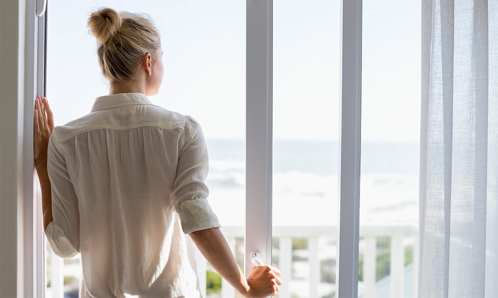 Eine Frau steht vor einem Fenster mit Blick auf das Meer, was die Wichtigkeit von guter Belüftung für Schimmelsanierung symbolisieren könnte.