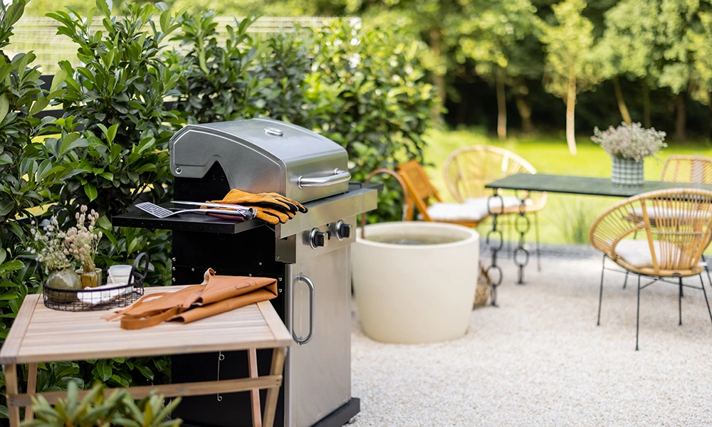 A modern grill with grill accessories on a wooden worktop, surrounded by lush greenery and a relaxing seating area in the background.