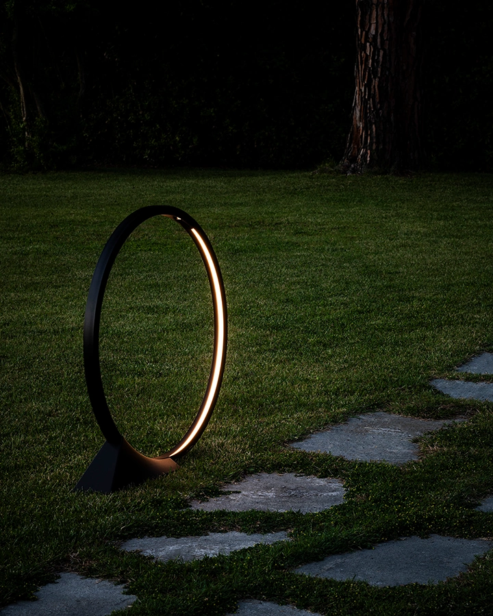 Ein ringförmiges Lichtobjekt erhellt einen Gartenpfad bei Nacht, das warme Licht schafft einen scharfen Kontrast zum dunklen Hintergrund.