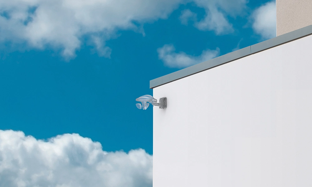 The image shows an outdoor light fixture mounted on the corner of a modern building's exterior wall. The sky is clear with scattered clouds, and the fixture has a minimalist, translucent design, adding a sleek look to the structure.